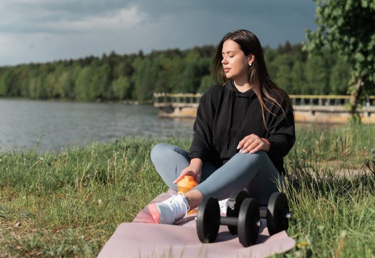 Woman sitting on yoga mat beside a lake with exercise equipment, enjoying a calm outdoor workout.