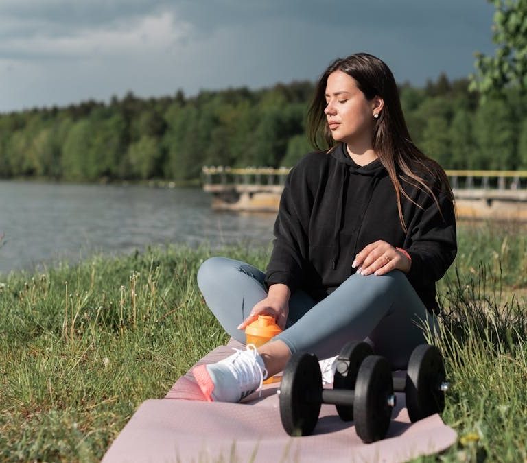 Woman sitting on yoga mat beside a lake with exercise equipment, enjoying a calm outdoor workout.